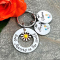 A silver metal keychain with hand stamped charms, including a large ring reading 'Best Mummy in the World' and smaller personalized names, displayed on a gray stone surface next to an orange rose.