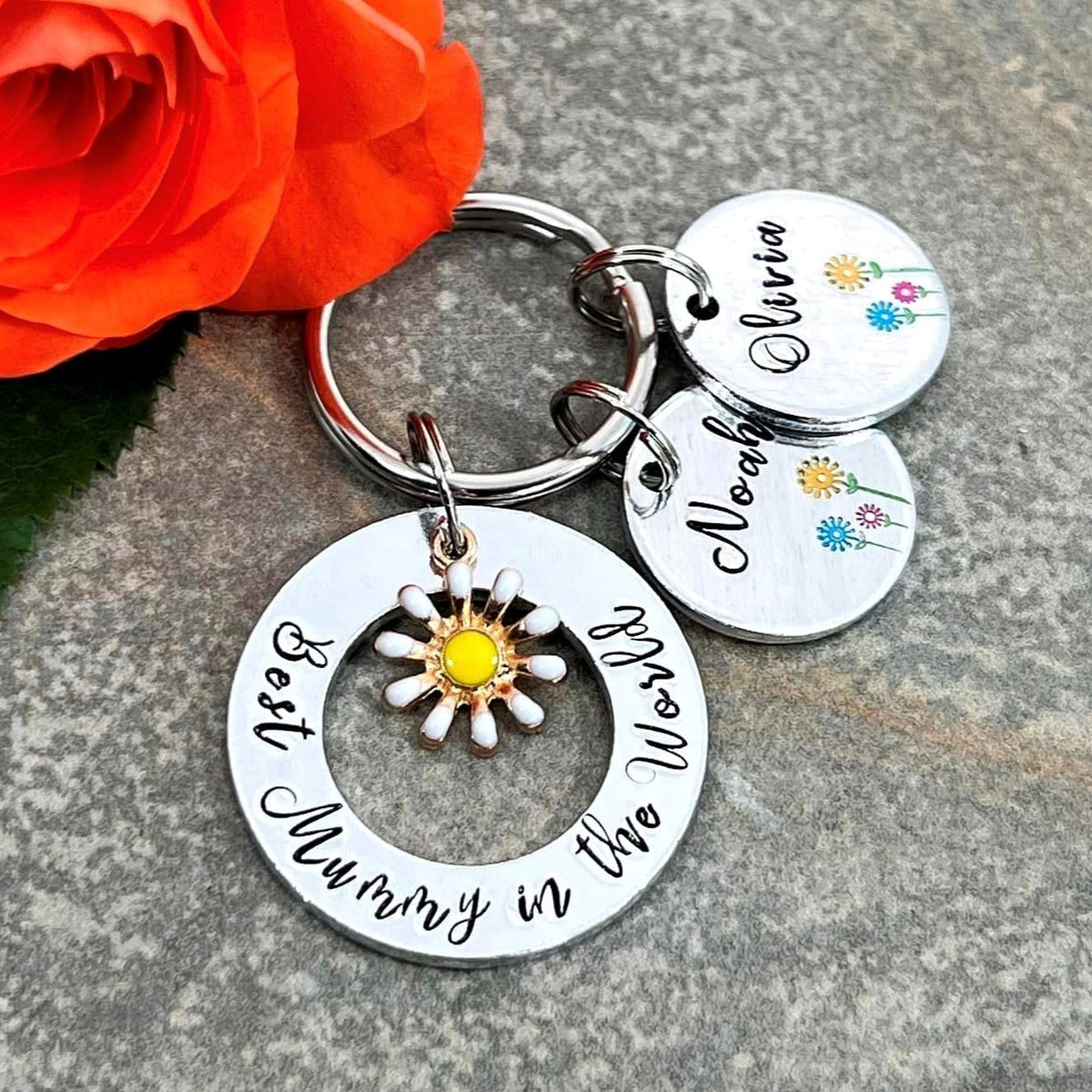A silver metal keychain with hand stamped charms, including a large ring reading 'Best Mummy in the World' and smaller personalized names, displayed on a gray stone surface next to an orange rose.
