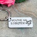 Hand-stamped You're My Lobster aluminium keyring with a lobster design on a grey stone surface beside a pink rose
