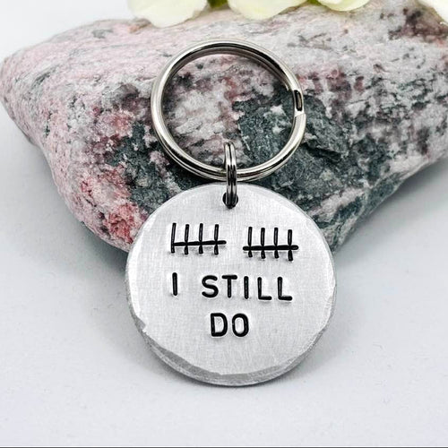 Aluminium I Still Do keyring with ten tally marks, displayed against a pink and grey stone with pale yellow flowers
