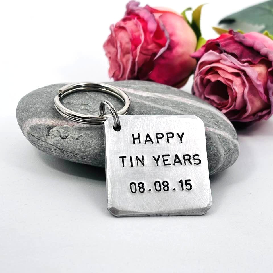 Personalised Happy Tin Years aluminium keyring resting against a grey stone, with pink roses in the background
