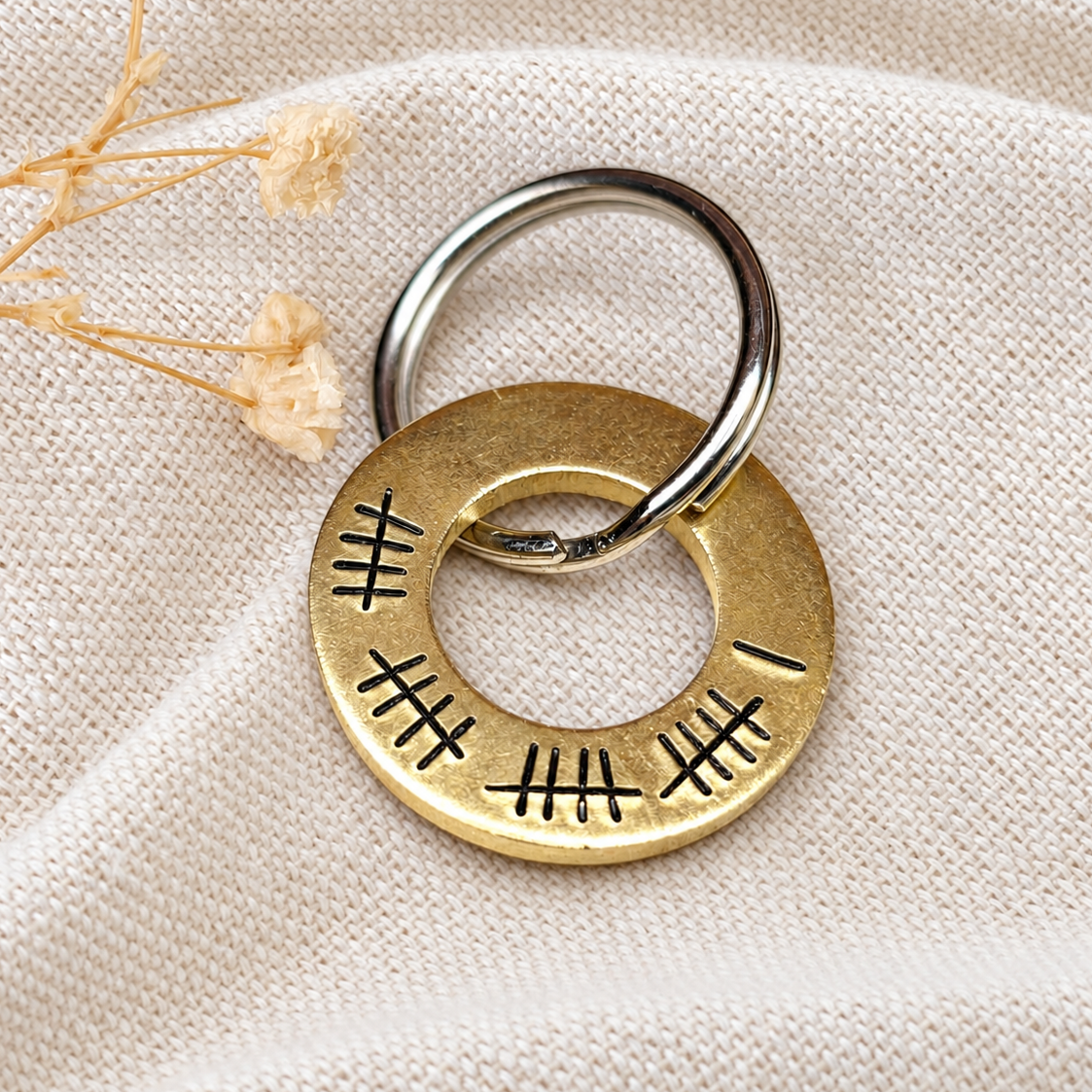 Brass 21st anniversary washer keyring with 21 hand stamped tally marks, displayed on linen beside dried flowers
