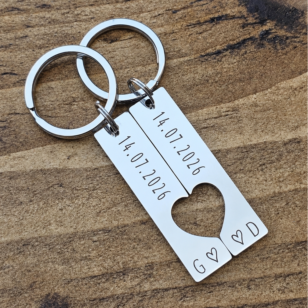 Two stainless steel date keyrings with a cut-out heart design displayed diagonally on a wooden surface
