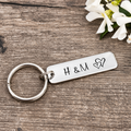 Personalised aluminium bar keyring stamped with H and M and two overlapping outline hearts on rustic wood beside white flowers
