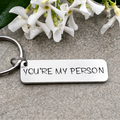 Close-up of the hand-stamped You're My Person lettering on an aluminium bar keyring beneath white flowers

