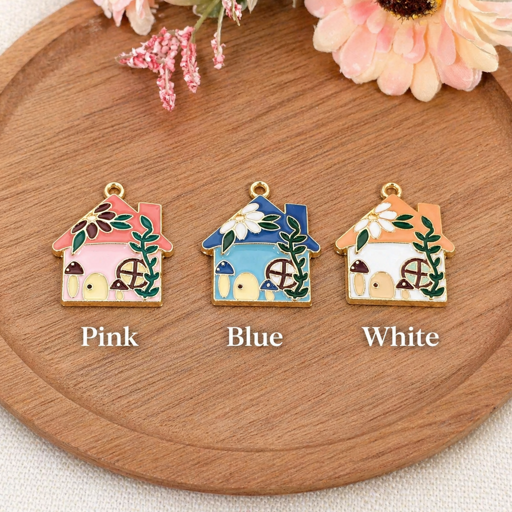 Three small house-shaped jewelry charms are displayed on a round wooden tray with flowers at the top. The charms are labeled Pink, Blue, and White underneath, showing different enamel colors.