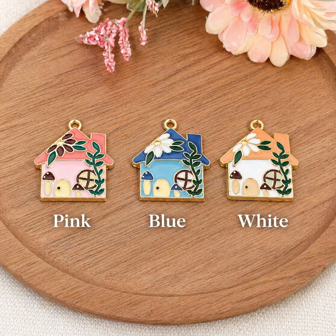 Three small house-shaped jewelry charms are displayed on a round wooden tray with flowers at the top. The charms are labeled Pink, Blue, and White underneath, showing different enamel colors.