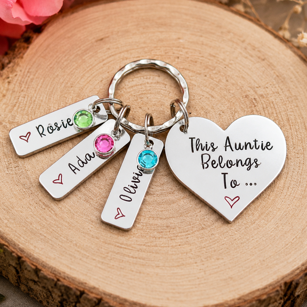 A silver metal keychain with four charms resting on a round wood slice. The keychain includes a heart-shaped charm that reads 'This Auntie Belongs To .
