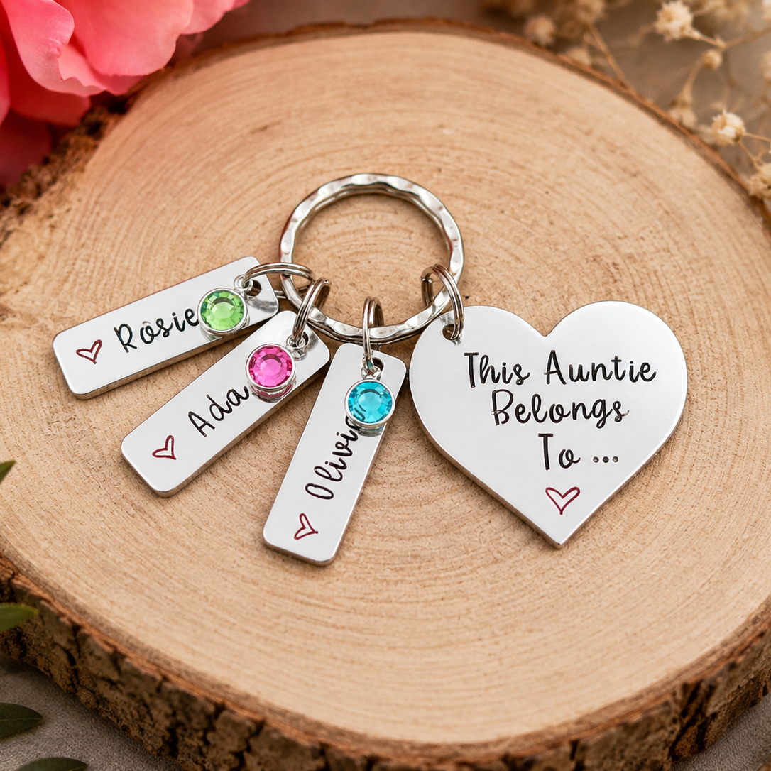 A silver metal keychain with four charms resting on a round wood slice. The keychain includes a heart-shaped charm that reads 'This Auntie Belongs To .