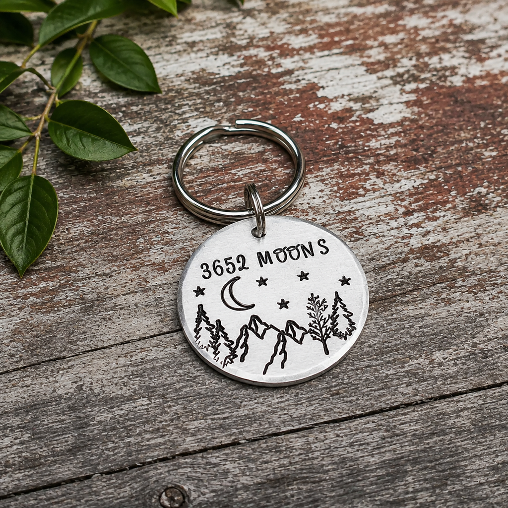 Round aluminium 3652 Moons keyring with hand stamped mountains, trees and stars on a weathered wooden surface
