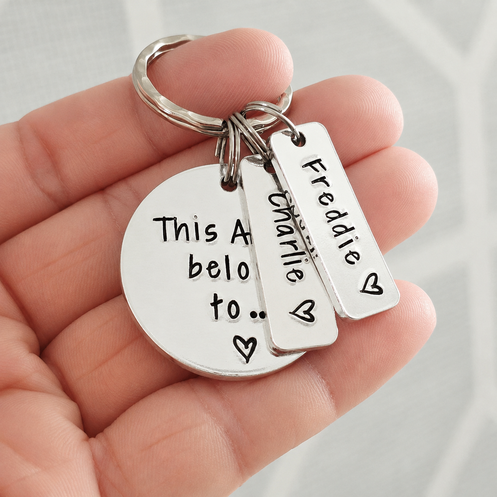 A silver metal keychain held in a hand, featuring a round disc charm with the text 'This A belo to.