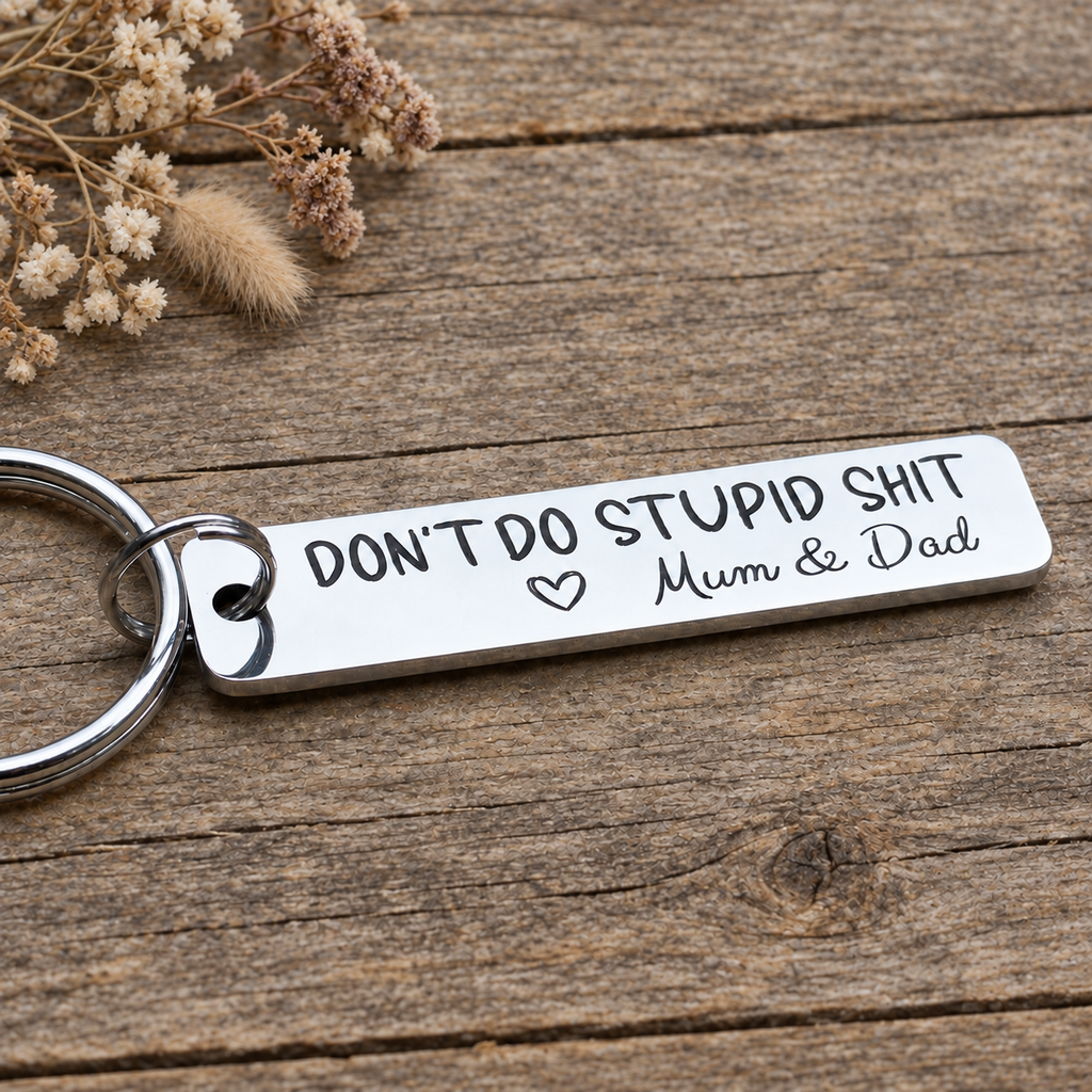 Angled view of an engraved Mum & Dad Don't Do Stupid Shit keyring on wood beside dried flowers