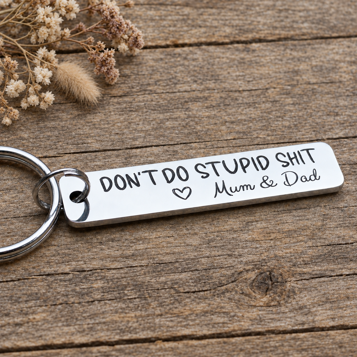 Angled view of an engraved Mum & Dad Don't Do Stupid Shit keyring on wood beside dried flowers