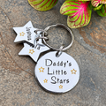 A silver metal keychain with a round disc engraved 'Daddy's Little Stars' and two star-shaped charms engraved with names, shown on a stone surface with decorative green leaves.