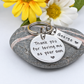 A silver-toned metal keychain set resting on a smooth gray stone, with yellow and white daisies in the background. The keychain features a round engraved disc reading 'Thank you for loving me as your own' and a smaller rectangular bar engraved with 'George' and a small heart.