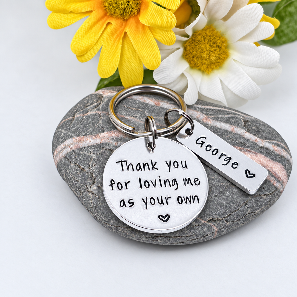 A silver-toned metal keychain with a round engraved disc reading 'Thank you for loving me as your own' and a smaller rectangular tag engraved 'George', displayed on a smooth gray stone with yellow and white flowers in the background.