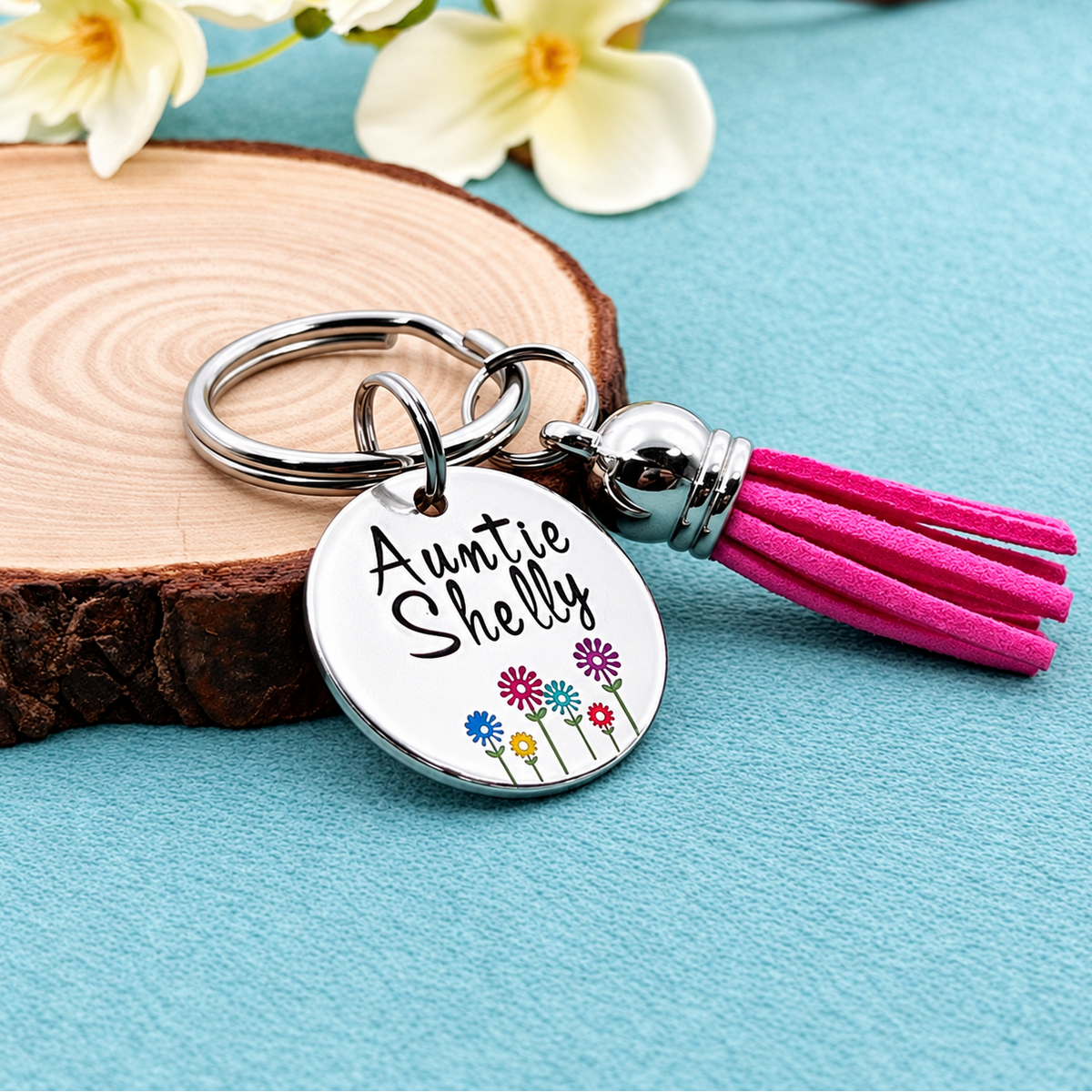 A personalized silver keychain with a round hand stamped disc reading 'Auntie Shelly' and small colorful flowers, attached to a bright pink tassel, styled on a blue surface with a wood slice and white flowers.