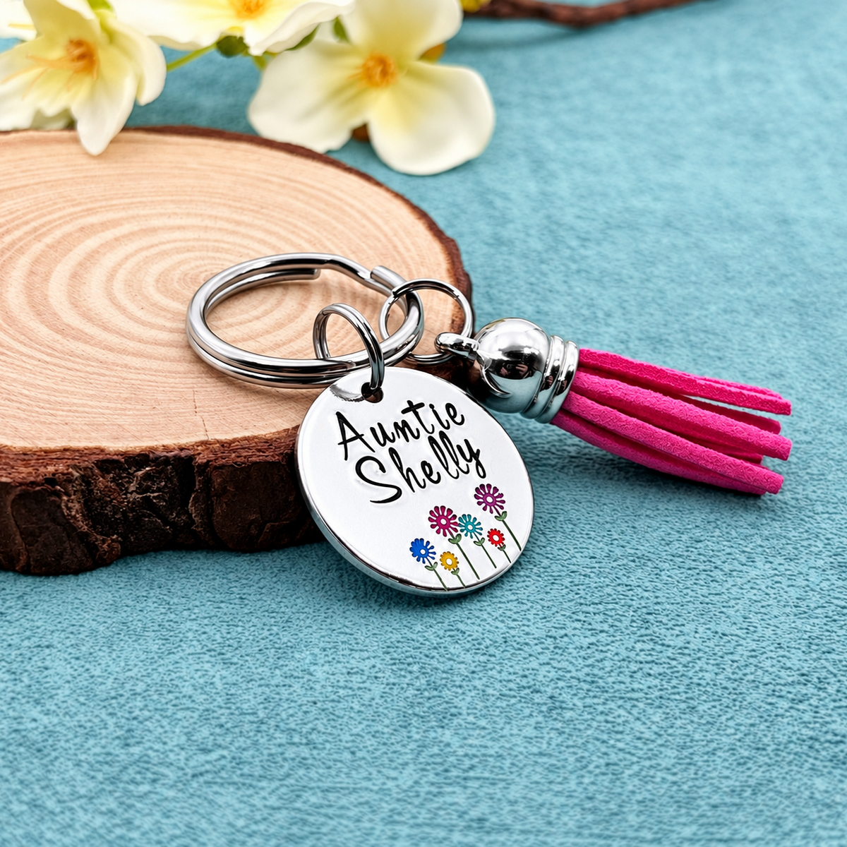 A personalized silver keychain with a round engraved disc reading 'Auntie Shelly' and small colorful flowers, attached to a bright pink tassel, styled on a blue textured surface with a wood slice and white flowers.