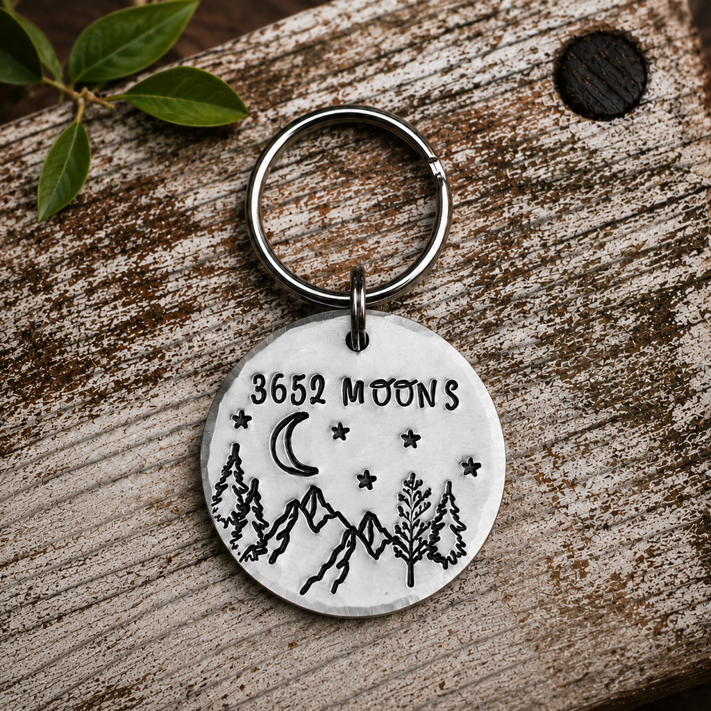 Aluminium 3652 Moons keyring with mountains, trees, a crescent moon and stars, displayed on rustic wood beside green leaves

