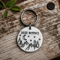 Aluminium 3652 Moons keyring with mountains, trees, a crescent moon and stars, displayed on rustic wood beside green leaves
