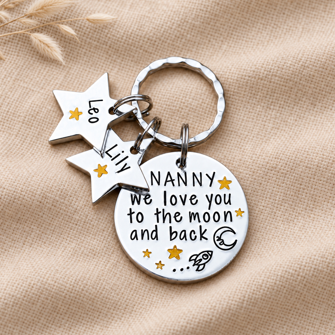 Personalised Nanny moon and back keyring with two star name tags, displayed on woven fabric beside dried stems