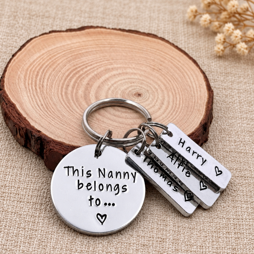 A silver metal keychain set with a round disc and three rectangular tags engraved with names and heart shapes, displayed on a rustic wood slice over a beige fabric background.