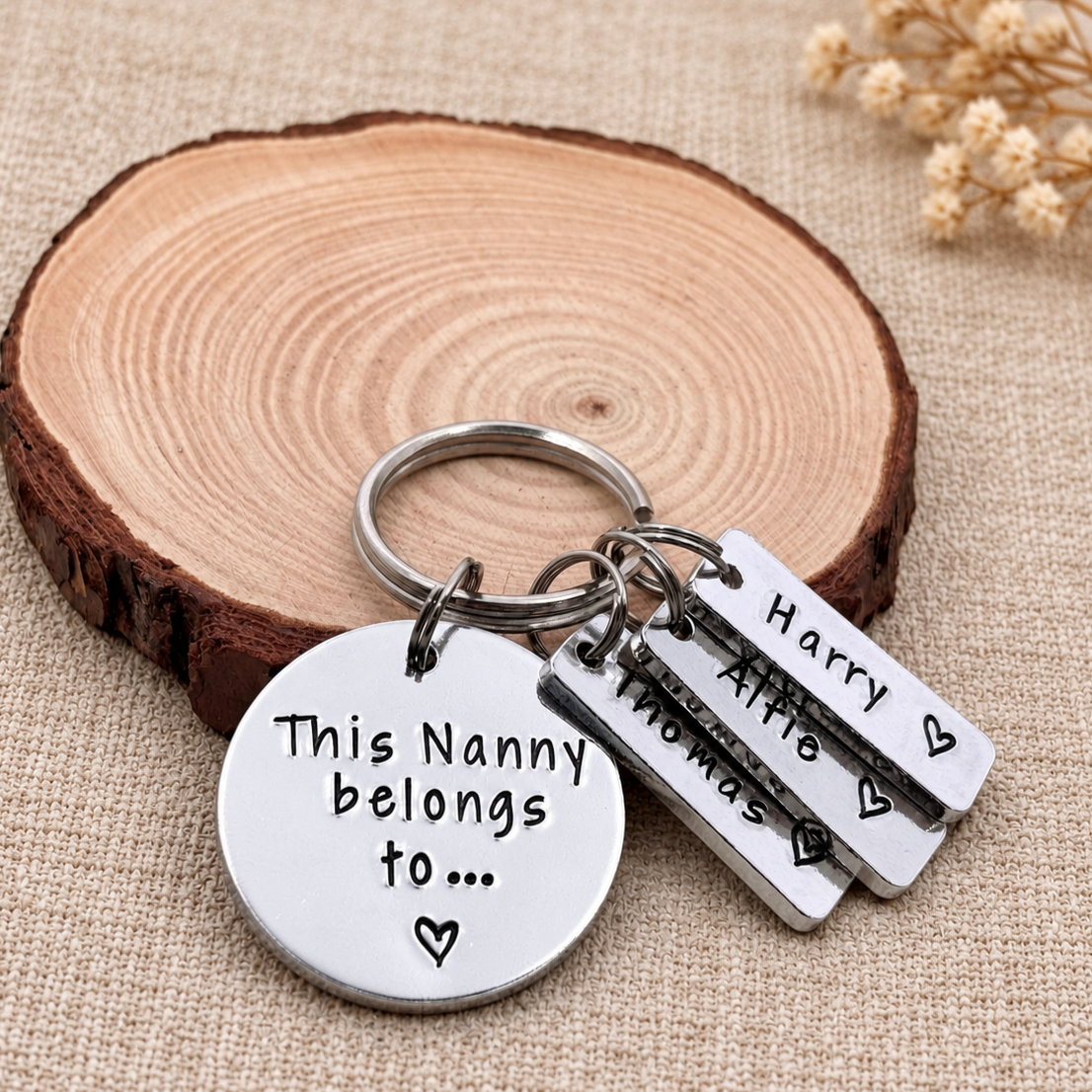 A silver metal keychain set with a round disc and three rectangular tags engraved with names and heart shapes, displayed on a rustic wood slice over a beige fabric background.
