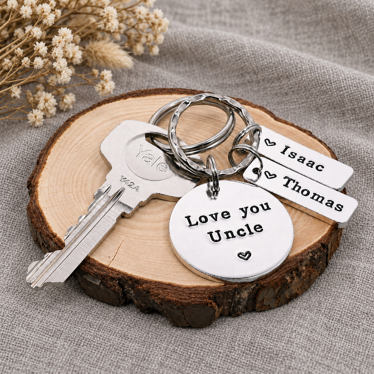 A silver metal keychain set displayed on a round wood slice coaster. The set includes a key, a round disc charm engraved with 'Love you Uncle' and a heart, two rectangular tags engraved with the names 'Isaac' and 'Thomas' next to a heart, and a silver keyring.