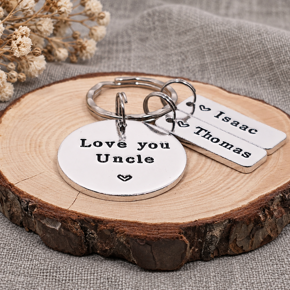 Personalized silver metal keychains displayed on a rustic wood slice. The set includes a round disc stamped with 'Love you Uncle' and a small heart, plus two rectangular tags engraved with the names 'Isaac' and 'Thomas'.