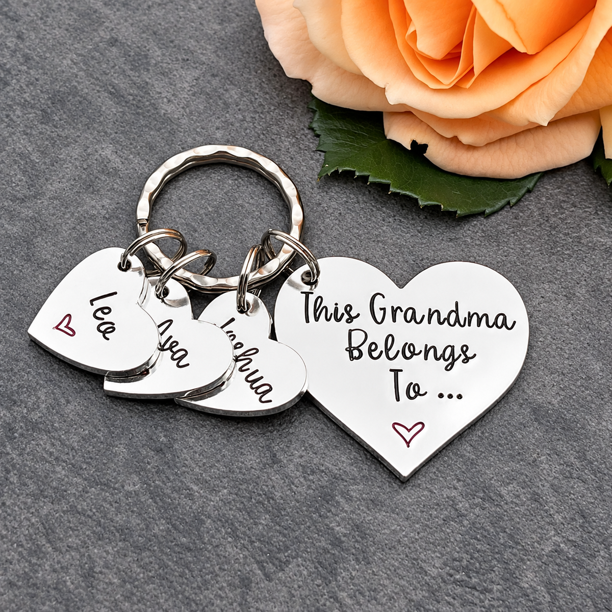 A silver heart-shaped keychain with three dangling heart charms on a dark gray textured surface, next to a peach rose. One large heart charm reads 'This Grandma Belongs To .