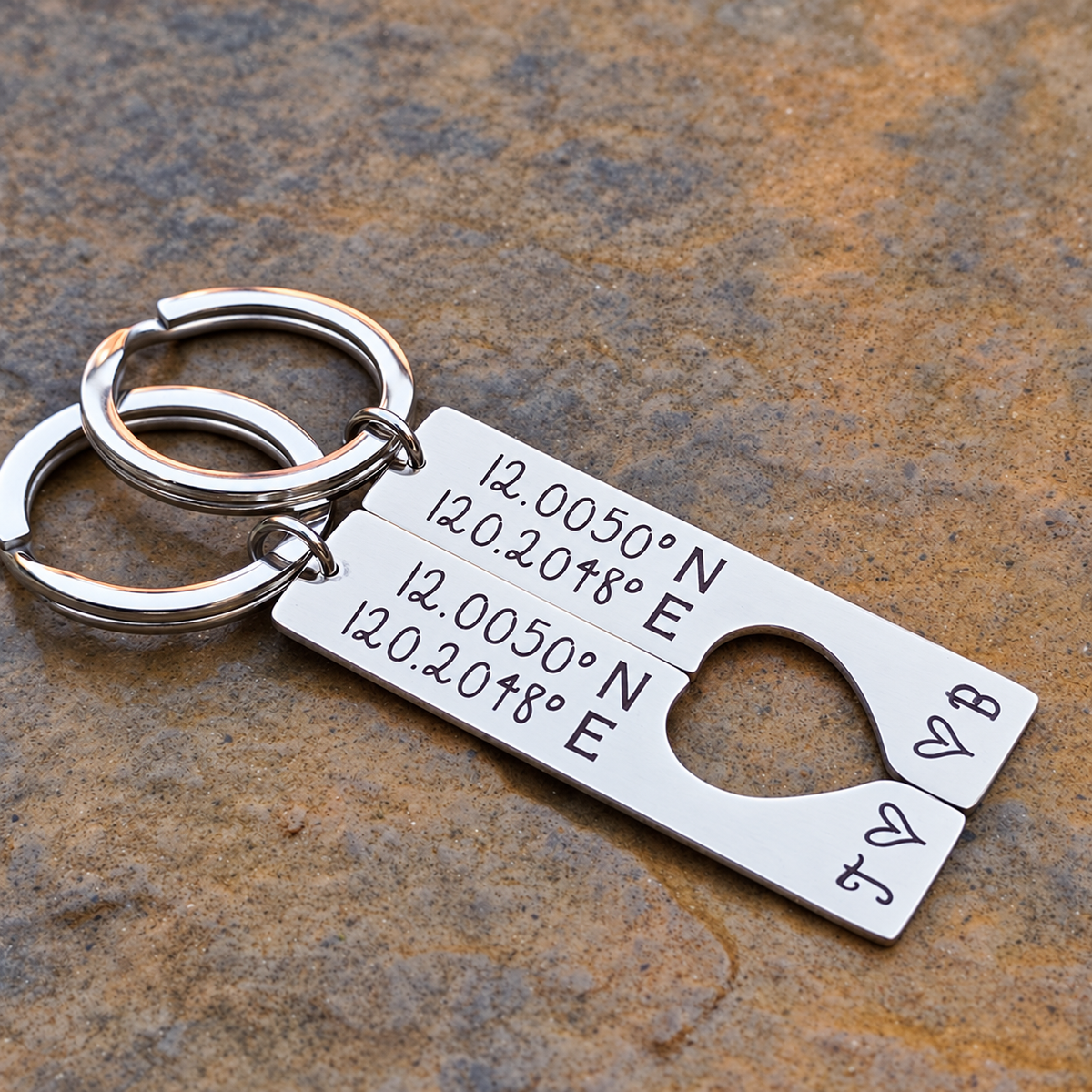 Angled close-up of matching engraved coordinates keyrings with initials J and B, small hearts and a heart-shaped cut-out on stone
