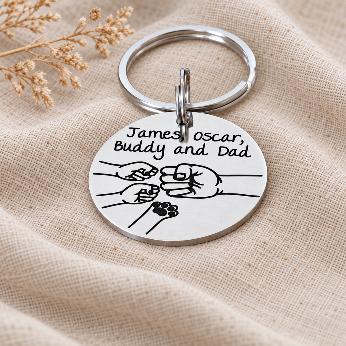 Close-up of an engraved family fist bump keyring showing personalised names, two children's fists and a pet paw beside Dad's fist
