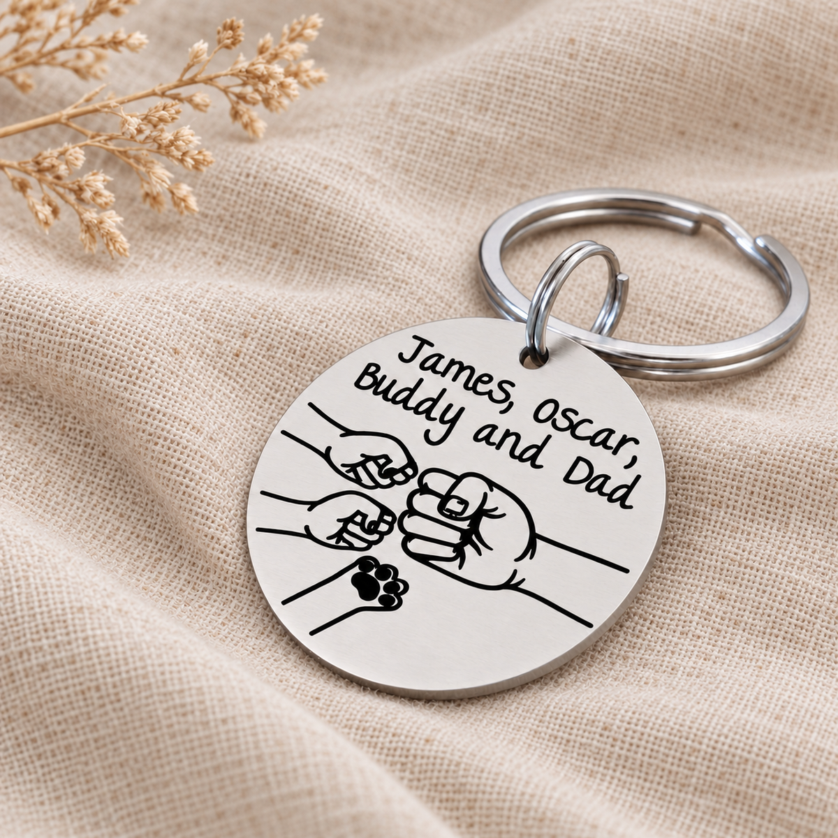 Angled view of a personalised Dad fist bump keyring with names and a pet paw design, resting on cream linen beside dried flowers
