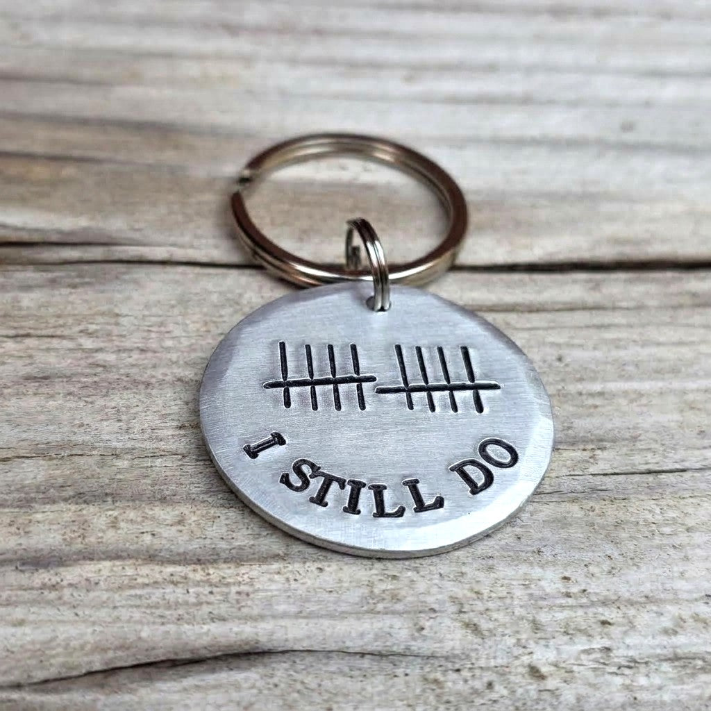 Angled view of a brushed aluminium I Still Do keyring with curved lettering on pale wooden boards

