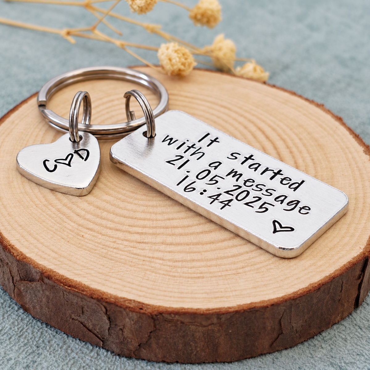 Angled view of a hand-stamped message keyring with a separate initial heart tag beside dried flowers
