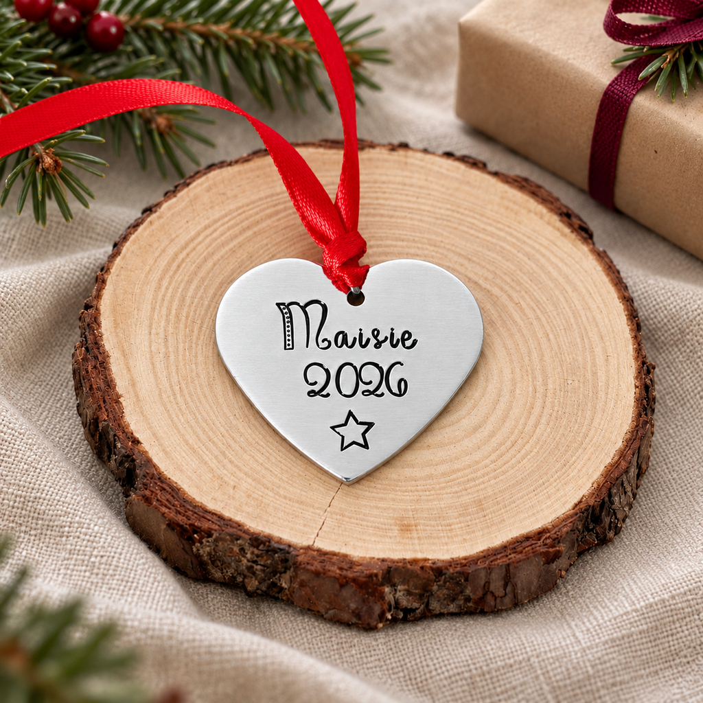 Personalised aluminium heart Christmas tree decoration stamped Maisie 2026 with a star and red ribbon, displayed upright on a wood slice
