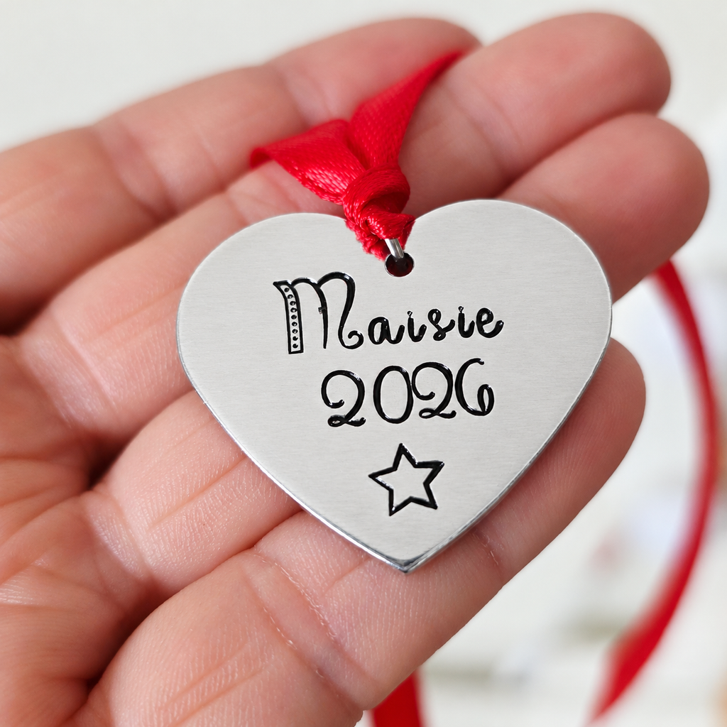 Personalised Maisie 2026 aluminium heart Christmas ornament held in a hand, showing the stamped star and red hanging ribbon

