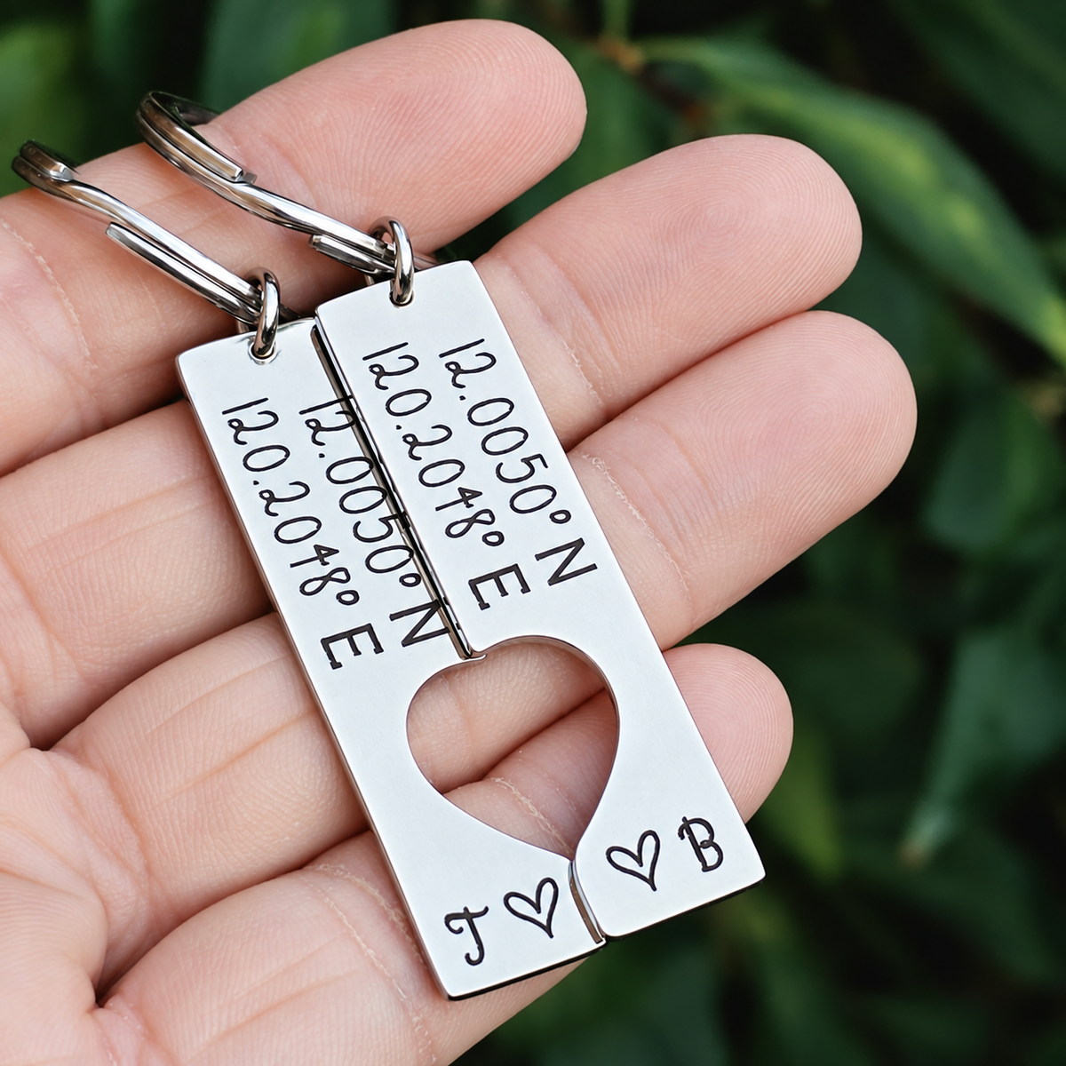 Personalised coordinates keyring set held in a hand, showing engraved stainless steel tags with initials J and B and a shared heart cut-out
