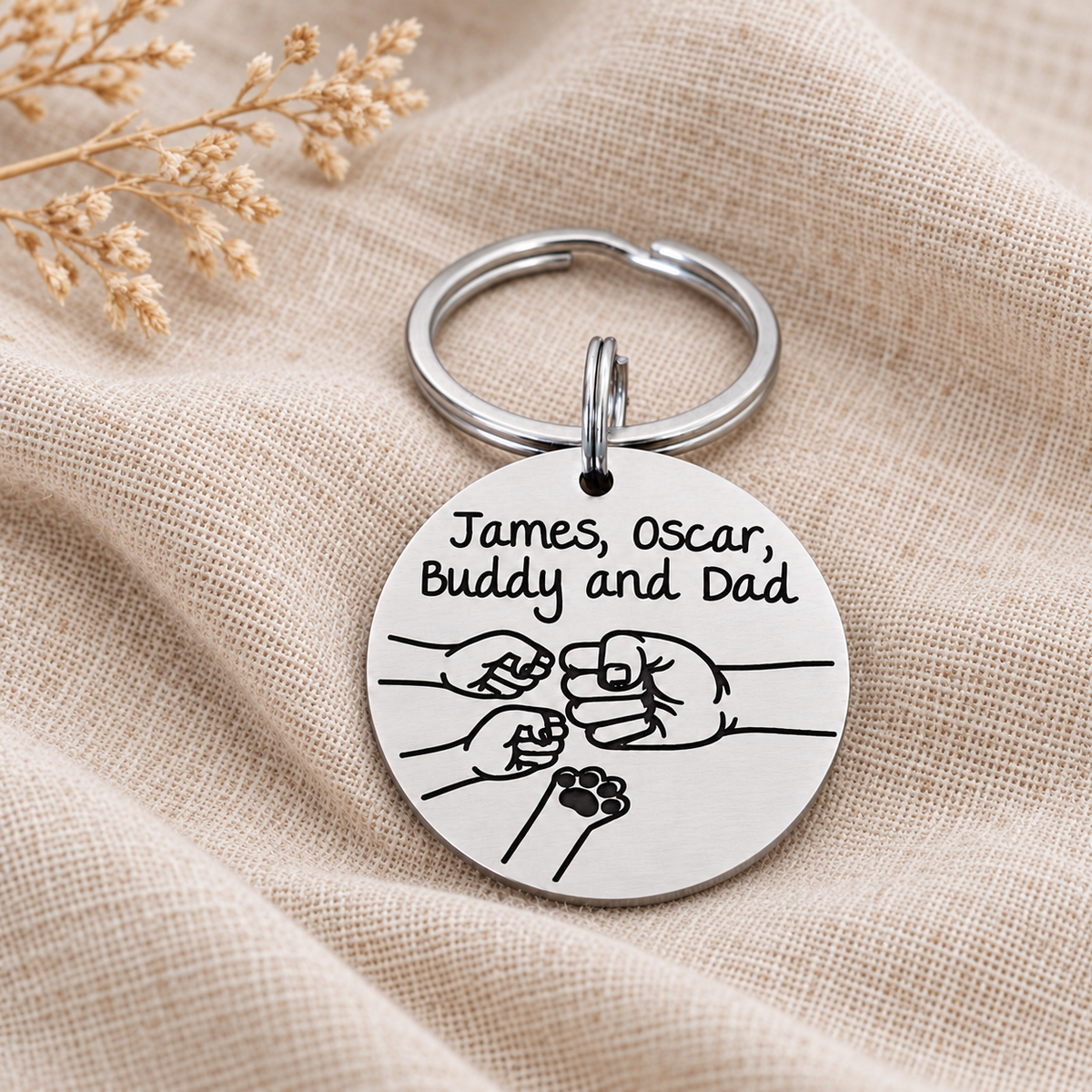 Round silver-coloured Dad keyring with personalised names, a family fist bump design and a pet paw, displayed on cream linen
