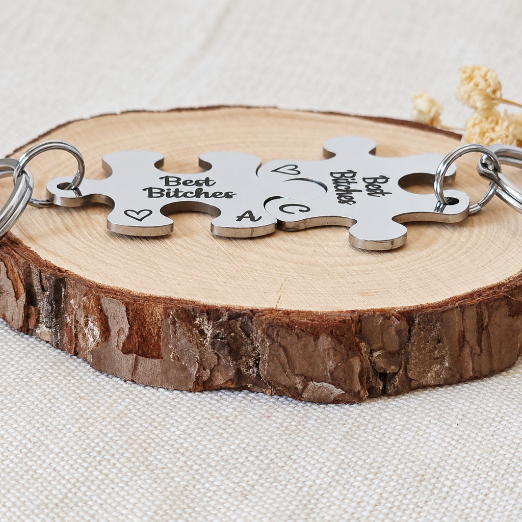 Low-angle view of two joined Best Bitches stainless steel puzzle keyrings on a wood slice
