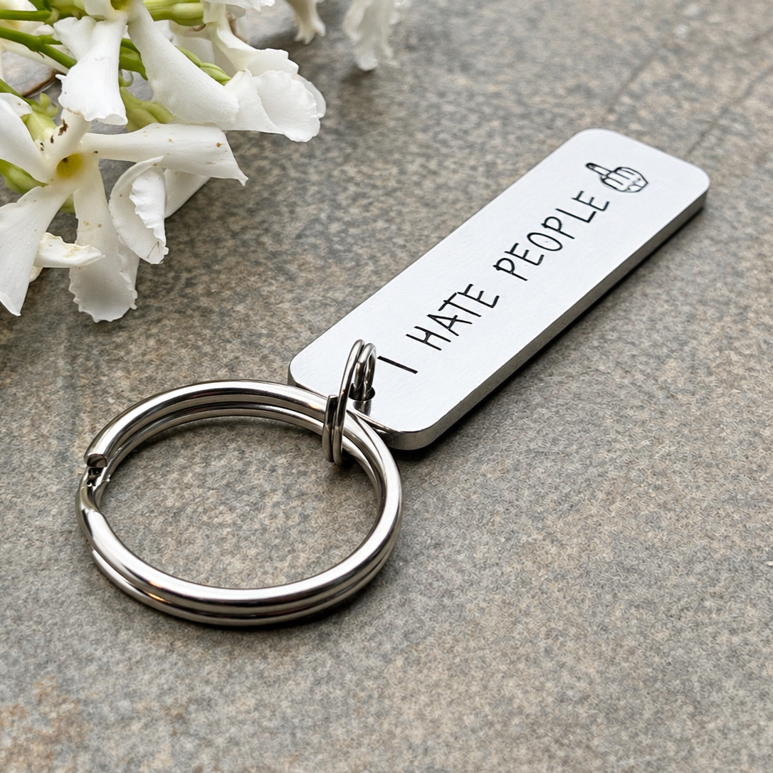 Low-angle view of an I Hate People keyring showing the aluminium edge and split ring beside white flowers
