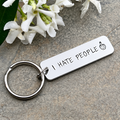 Hand-stamped aluminium I Hate People keyring with a middle finger design beside white flowers on a grey stone surface
