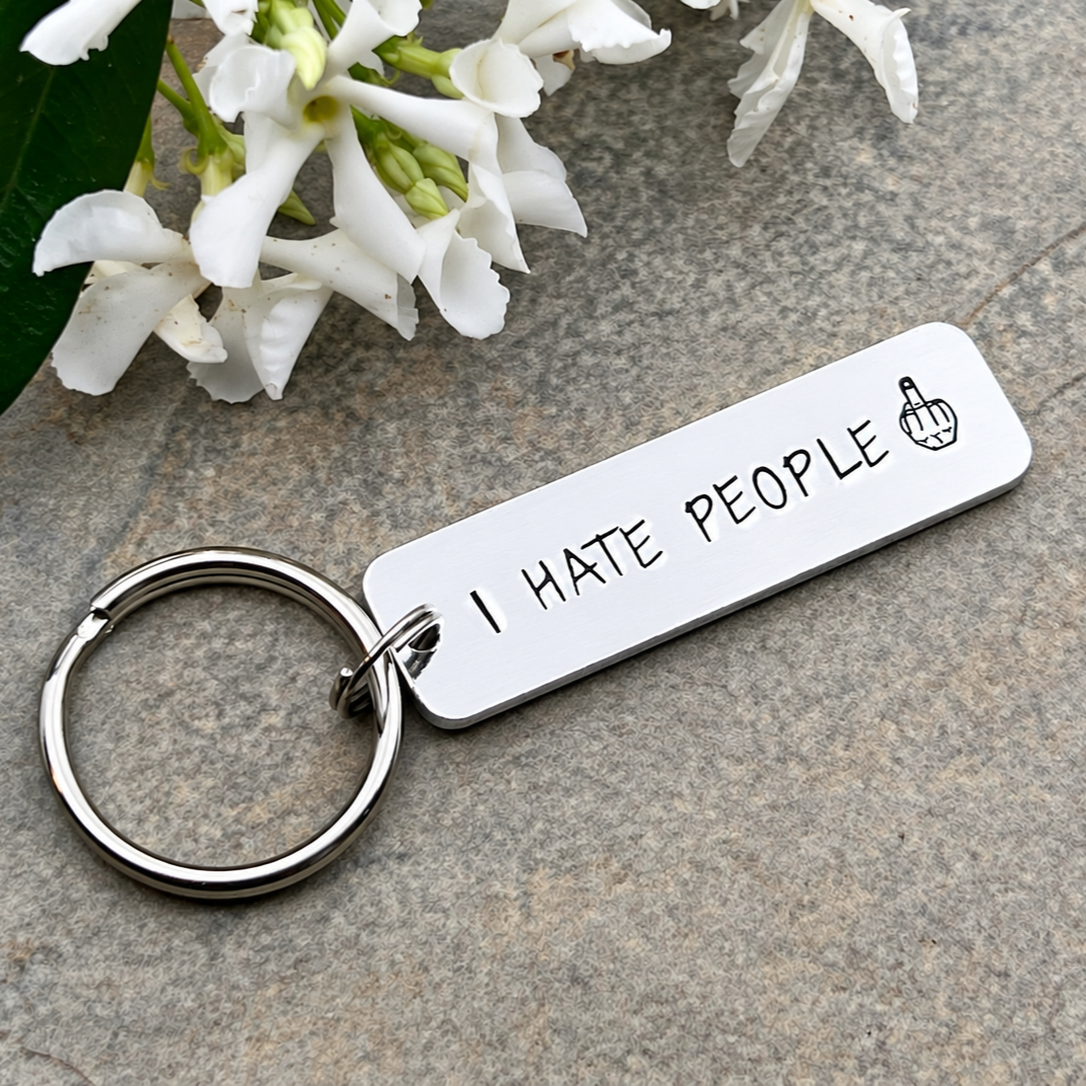 Hand-stamped aluminium I Hate People keyring with a middle finger design beside white flowers on a grey stone surface
