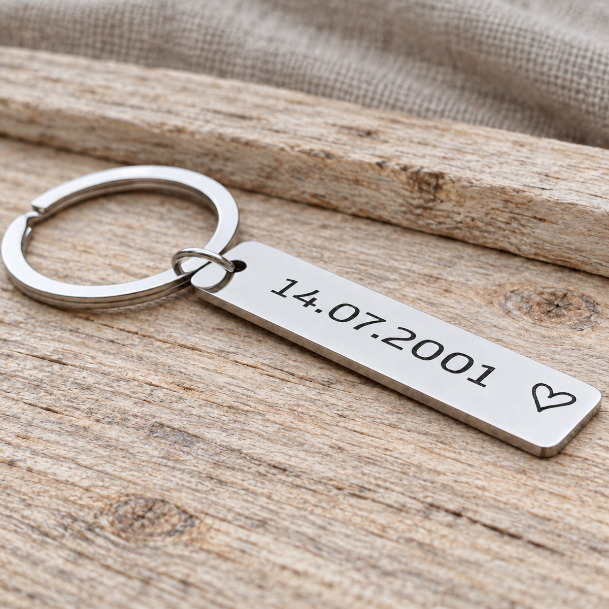 Angled close-up of a personalised date keyring with an outline heart on pale wood beside beige fabric
