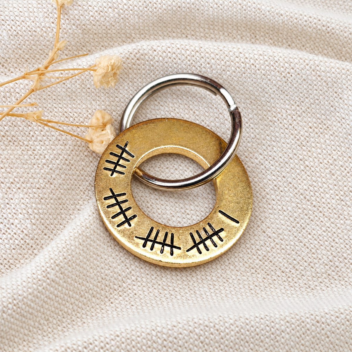Front view of a brass washer keyring with 21 tally marks, resting on folded linen beside dried flowers
