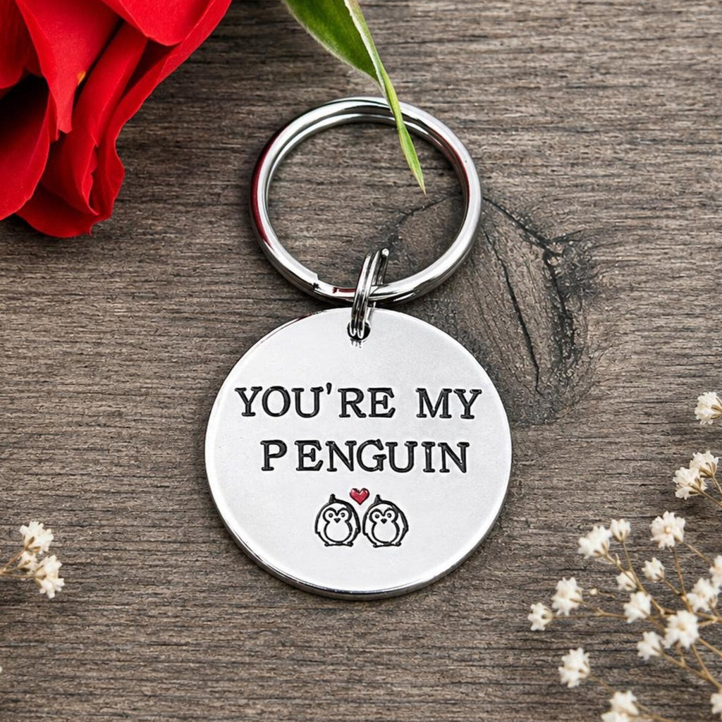 Round aluminium You're My Penguin keyring with two penguins and a red heart on rustic wood beside a red rose
